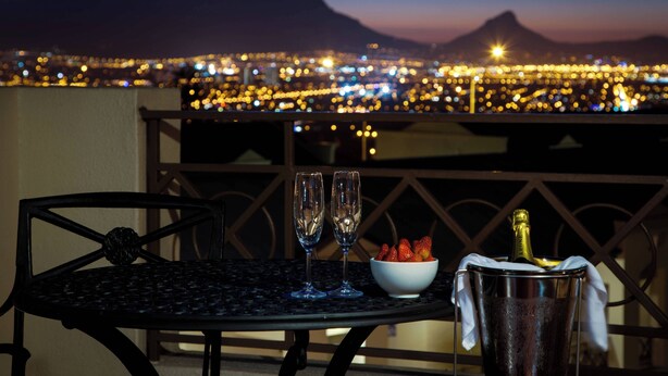 A table with two glasses, a bowl of strawberries, and a bottle of champagne on a balcony overlooking a cityscape at dusk.