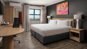 A hotel room with a large bed, a desk, a chair, a TV, a window with curtains, and a painting on the wall.