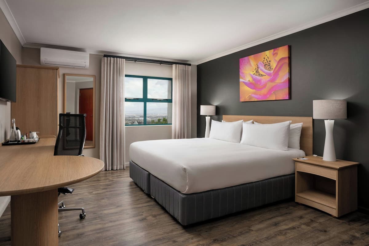 Ein Hotelzimmer mit einem großen Bett, einem Schreibtisch, einem Stuhl, einem Fernseher, einem Fenster mit Vorhängen und einem Bild an der Wand.