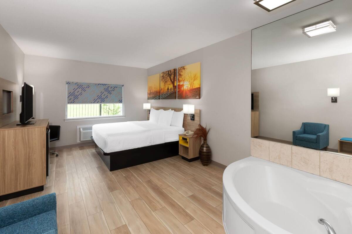 A modern hotel room with a large bed, a sitting area, a bathtub, and a television.