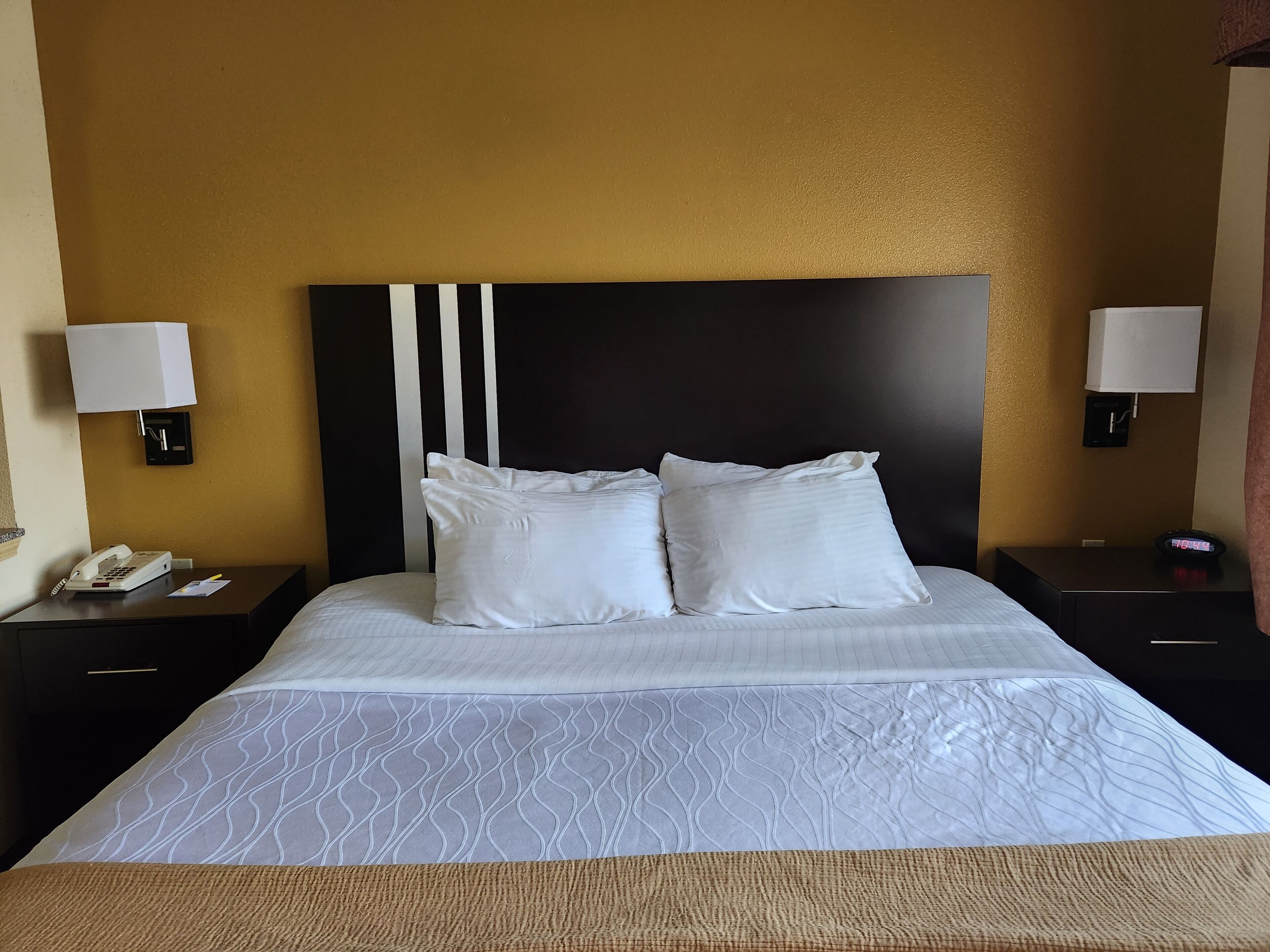 A hotel room with a large bed, two bedside tables, white lamps, a telephone, and a clock.