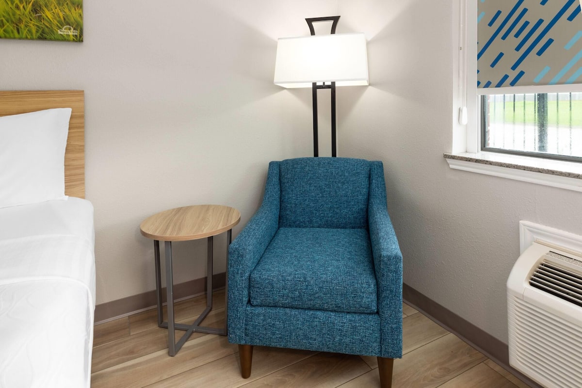 A room with a blue armchair, a small round table, a white bed, a wall-mounted lamp, and a window with blinds.