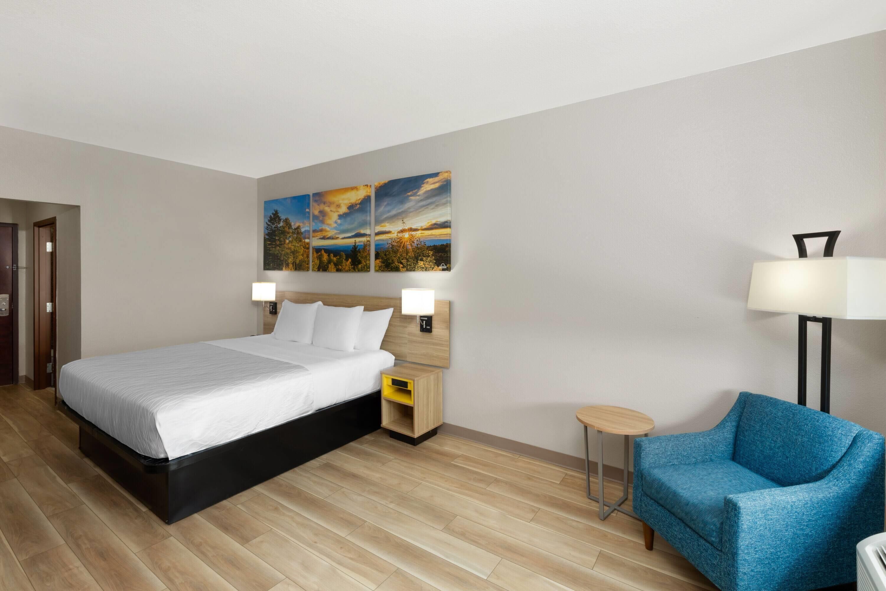Una habitación de hotel con una cama grande, un sillón azul, una mesita de noche y una lámpara de pared.