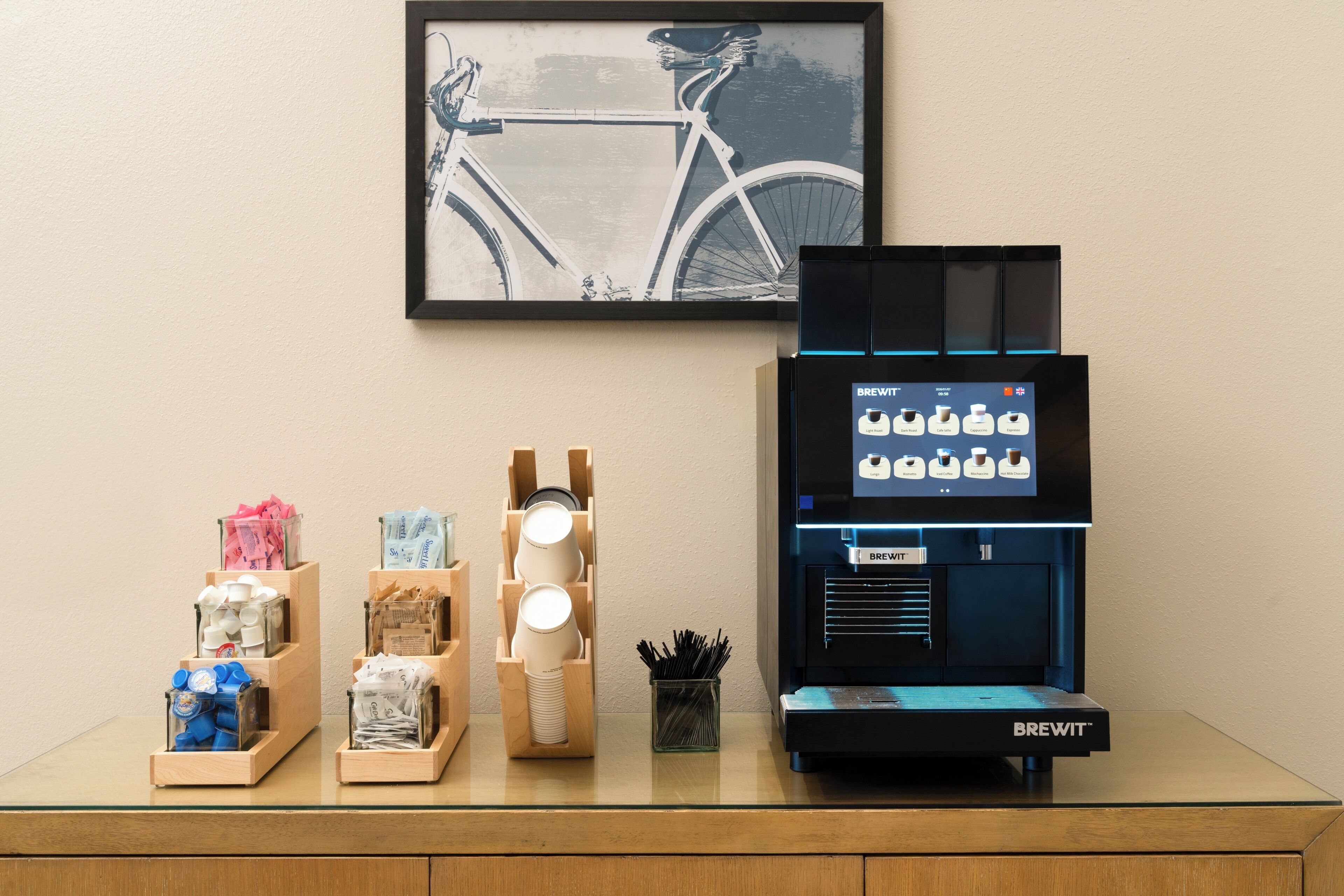 A coffee machine with a digital display, various coffee pods, and a framed bicycle artwork on a counter.