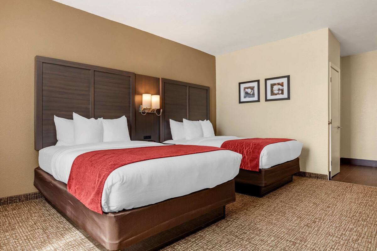 A hotel room with two beds, a large wooden headboard, a red throw blanket, and two framed pictures on the wall.