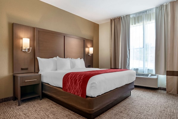 A hotel room with a large bed, two bedside tables, wall-mounted lamps, an air conditioning unit, and a window with curtains.