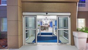 A hotel entrance with automatic sliding doors, a reception desk, and a blue carpeted hallway leading to rooms. - Holiday Inn Express New Orleans East by IHG (New Orleans)