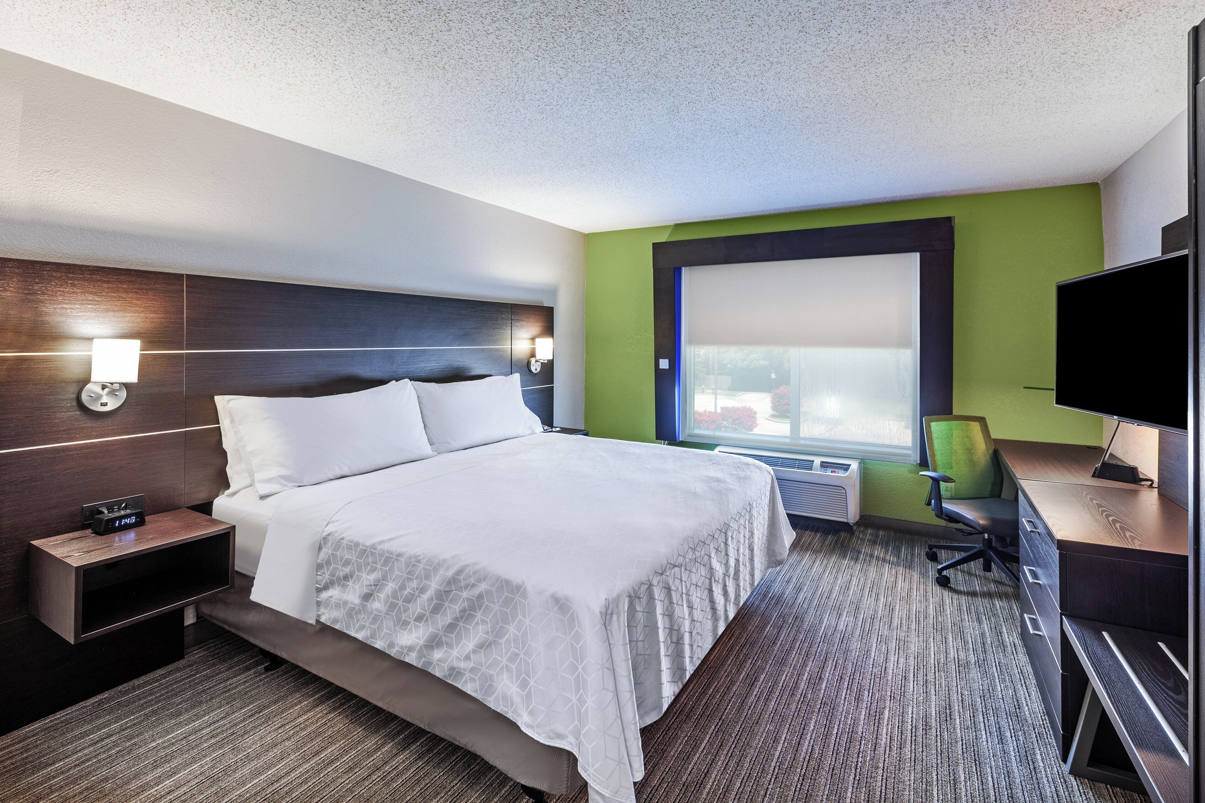 Een hotelkamer met een bed, nachtkastje met een klok, een bureau met stoel, een televisie en een raam.