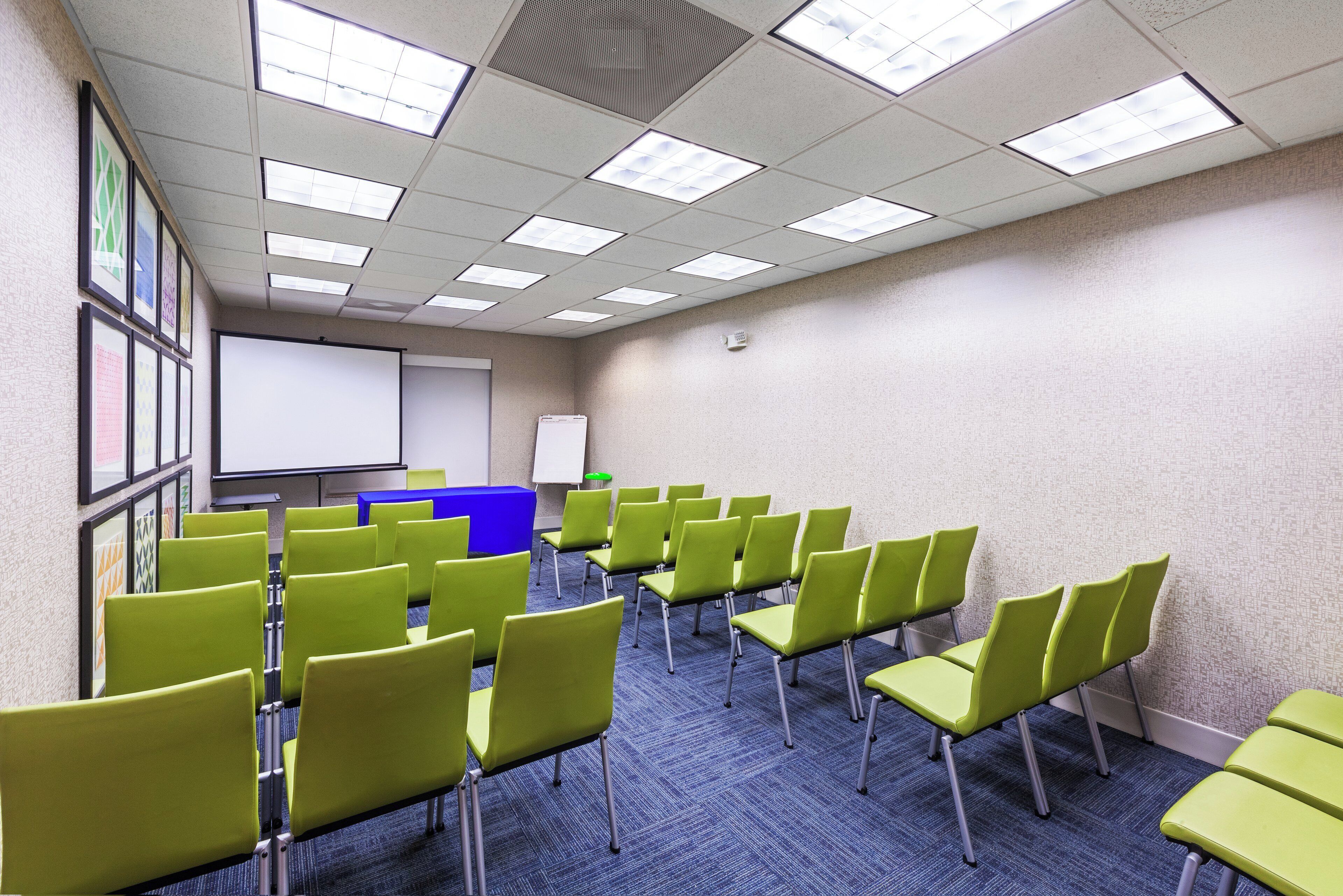 A conference room with rows of green chairs, a whiteboard, and a projector screen.