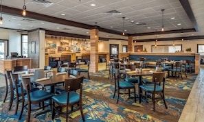 A hotel dining area with multiple tables and chairs, a counter with a menu, and artwork on the walls. - Holiday Inn Conference Ctr Marshfield by IHG (Marshfield)