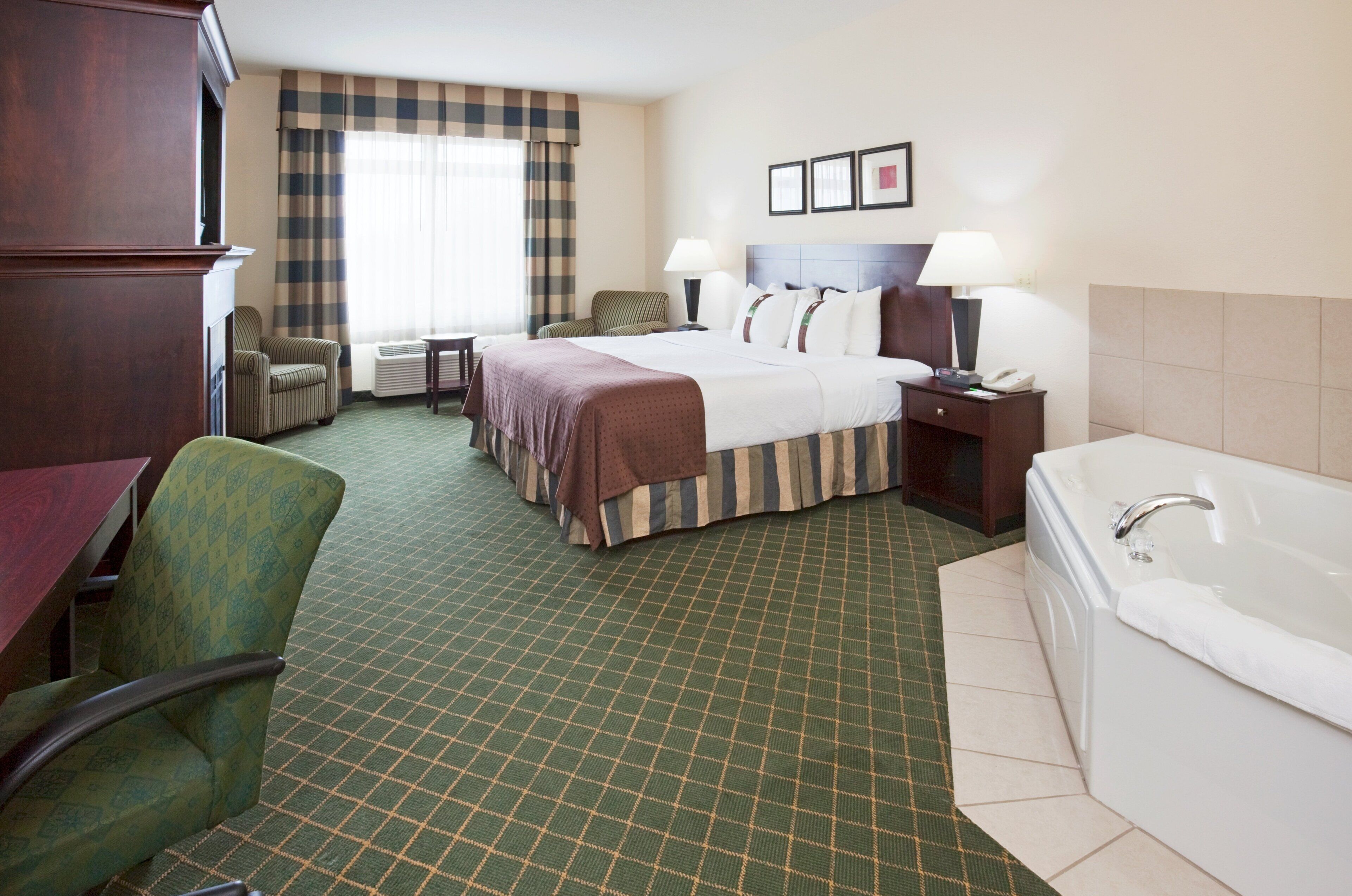 A hotel room with a bed, two bedside tables, a chair, a desk, a desk lamp, a telephone, a wall-mounted TV, and a bathroom with a bathtub and toilet.