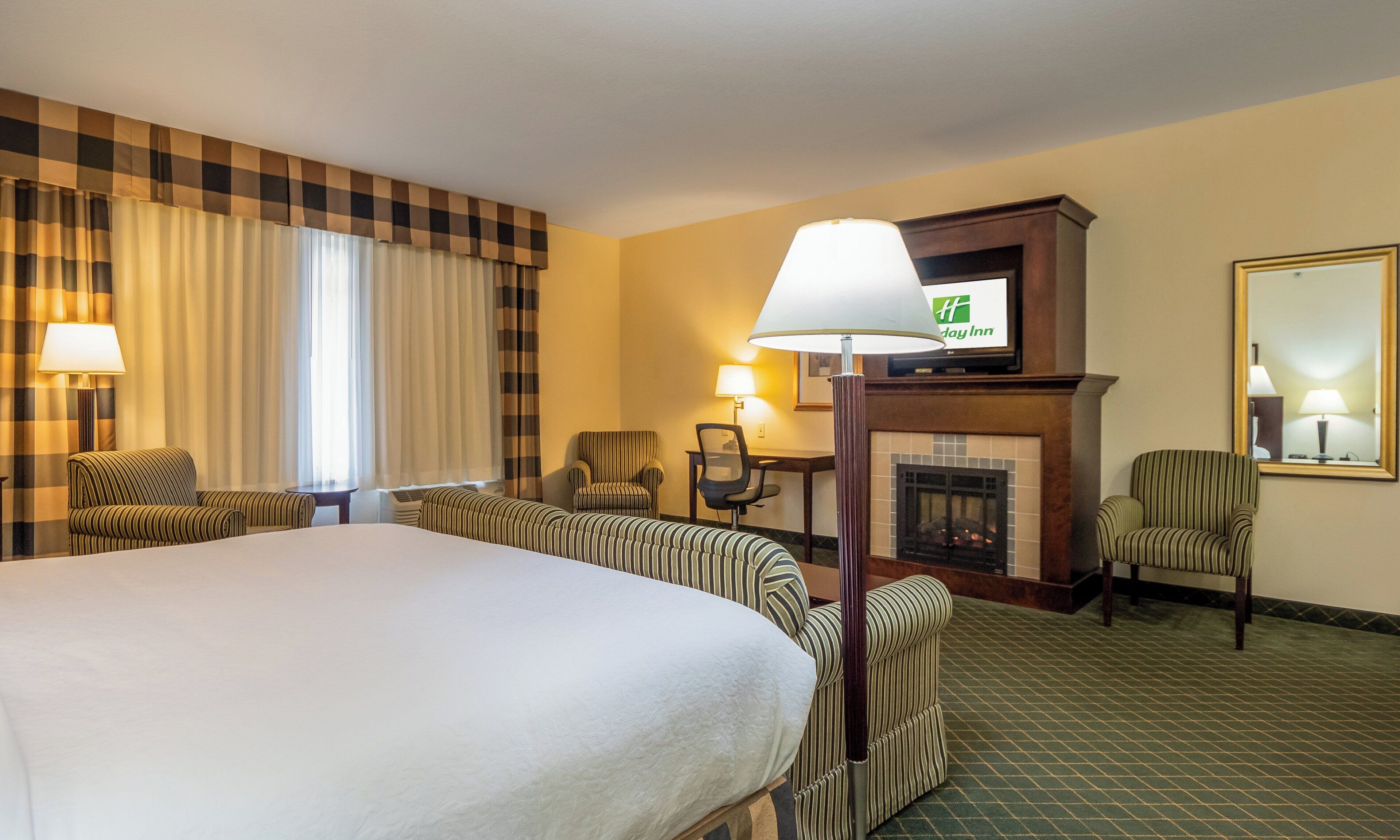A hotel room with a large bed, two armchairs, a desk, a fireplace, and a television on the wall.