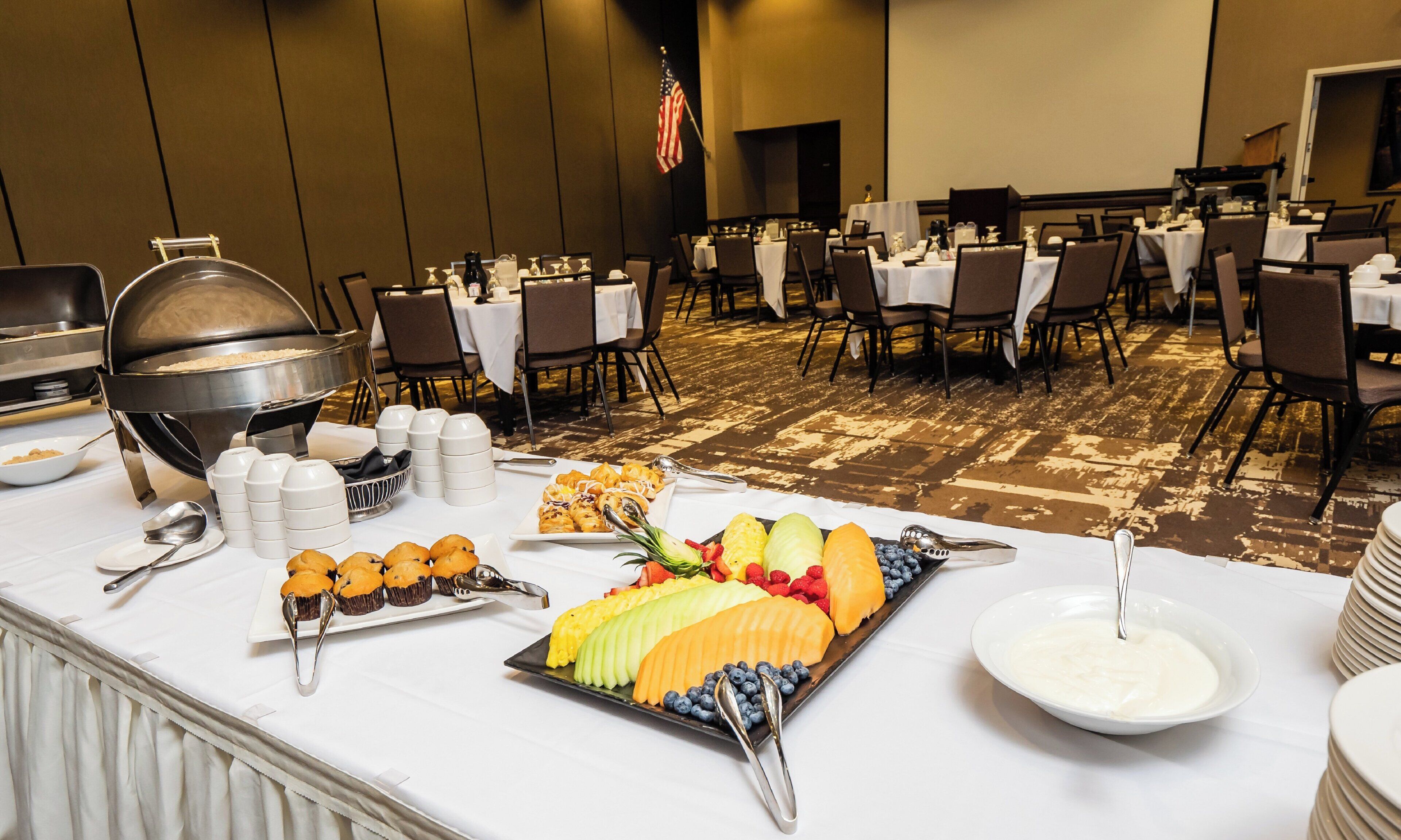 A buffet setup with various food items and utensils on a table, surrounded by chairs and tables in a banquet hall.
