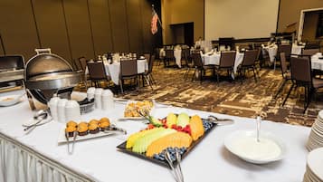 A buffet setup with various food items and utensils on a table, surrounded by chairs and tables in a banquet hall.