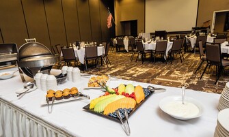 Montaje de bufé con diversos alimentos y utensilios sobre una mesa, rodeado de sillas y mesas en un salón de banquetes.