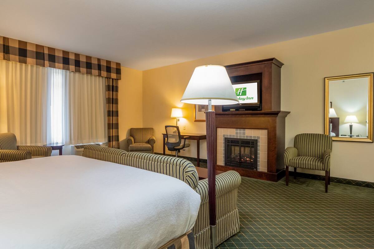 A hotel room with a large bed, two armchairs, a desk with a chair, a fireplace, and a television mounted above the fireplace.