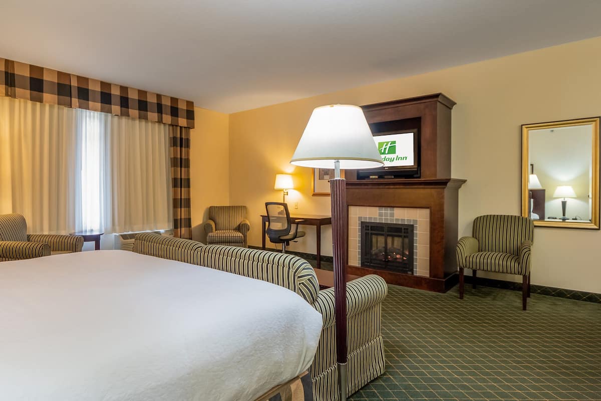 A hotel room with a large bed, two armchairs, a desk with a chair, a fireplace, and a television mounted above the fireplace.