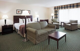 Una habitación de hotel con una cama, un sofá a rayas, una mesita, dos sillones y una mesita de noche con una lámpara.