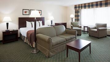Habitación de hotel con cama, sofá a rayas, una mesita, dos sillones, una mesita de noche con lámpara y ventana con cortinas.