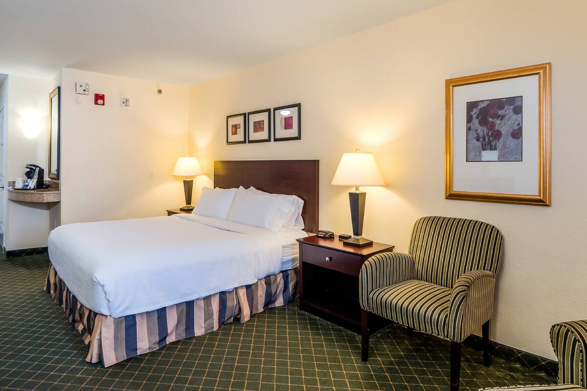 A hotel room with a bed, two bedside tables with lamps, a chair, a desk with a coffee maker, and framed pictures on the wall.