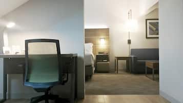 Habitación de hotel con escritorio, silla de oficina, cama, mesita de noche, lámpara y un cuadro en la pared.