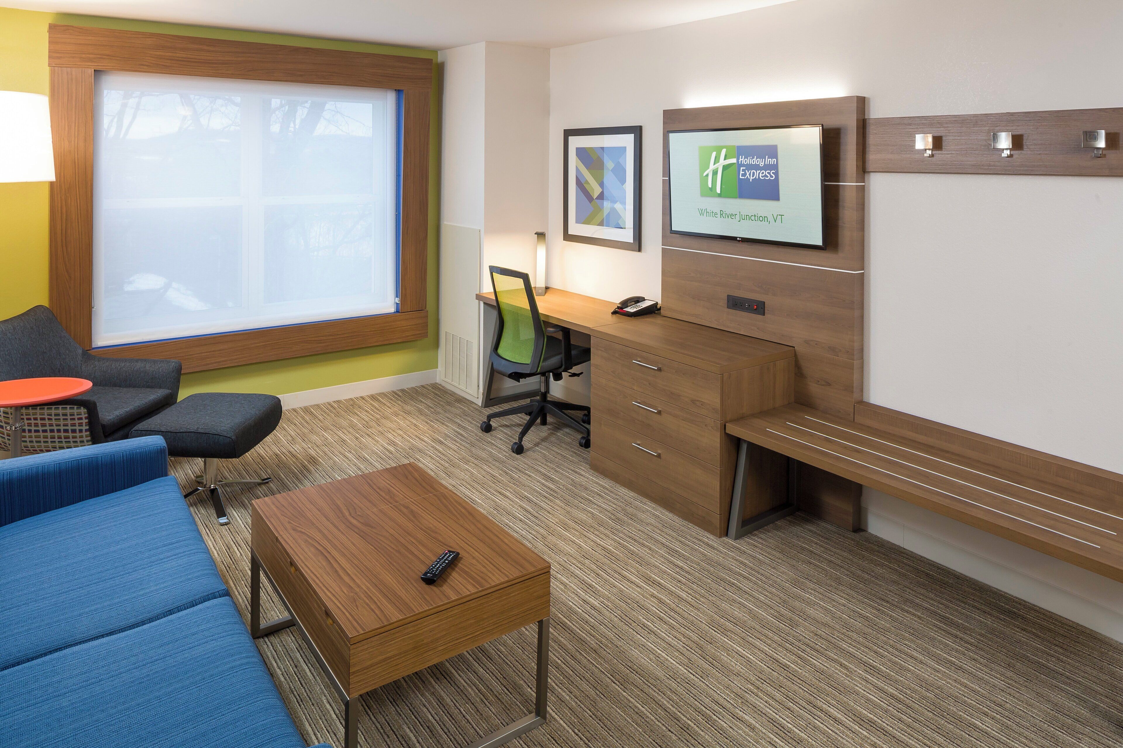 Una camera d'albergo con una scrivania, una sedia, un divano, un tavolino da caffè e uno schermo televisivo che mostra il logo Holiday Inn Express.