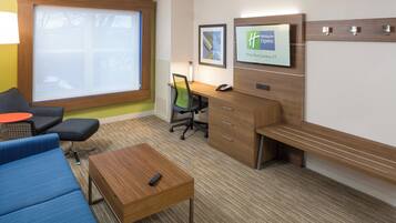 Una camera d'albergo con una scrivania, una sedia, un divano, un tavolino da caffè e uno schermo televisivo che mostra il logo Holiday Inn Express.