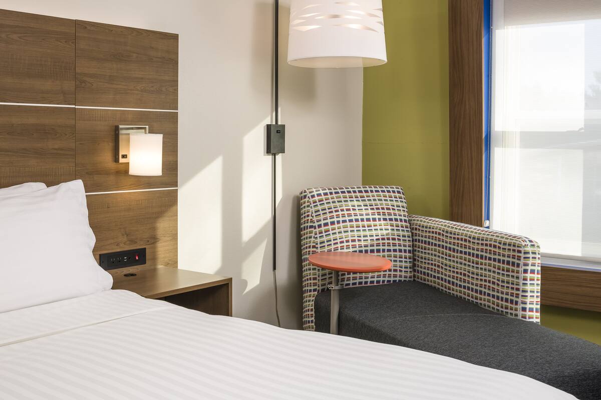 Une chambre d’hôtel avec un lit, une table de chevet, une chaise avec un coussin à motifs, une lampe fixée au mur et une fenêtre avec des stores.