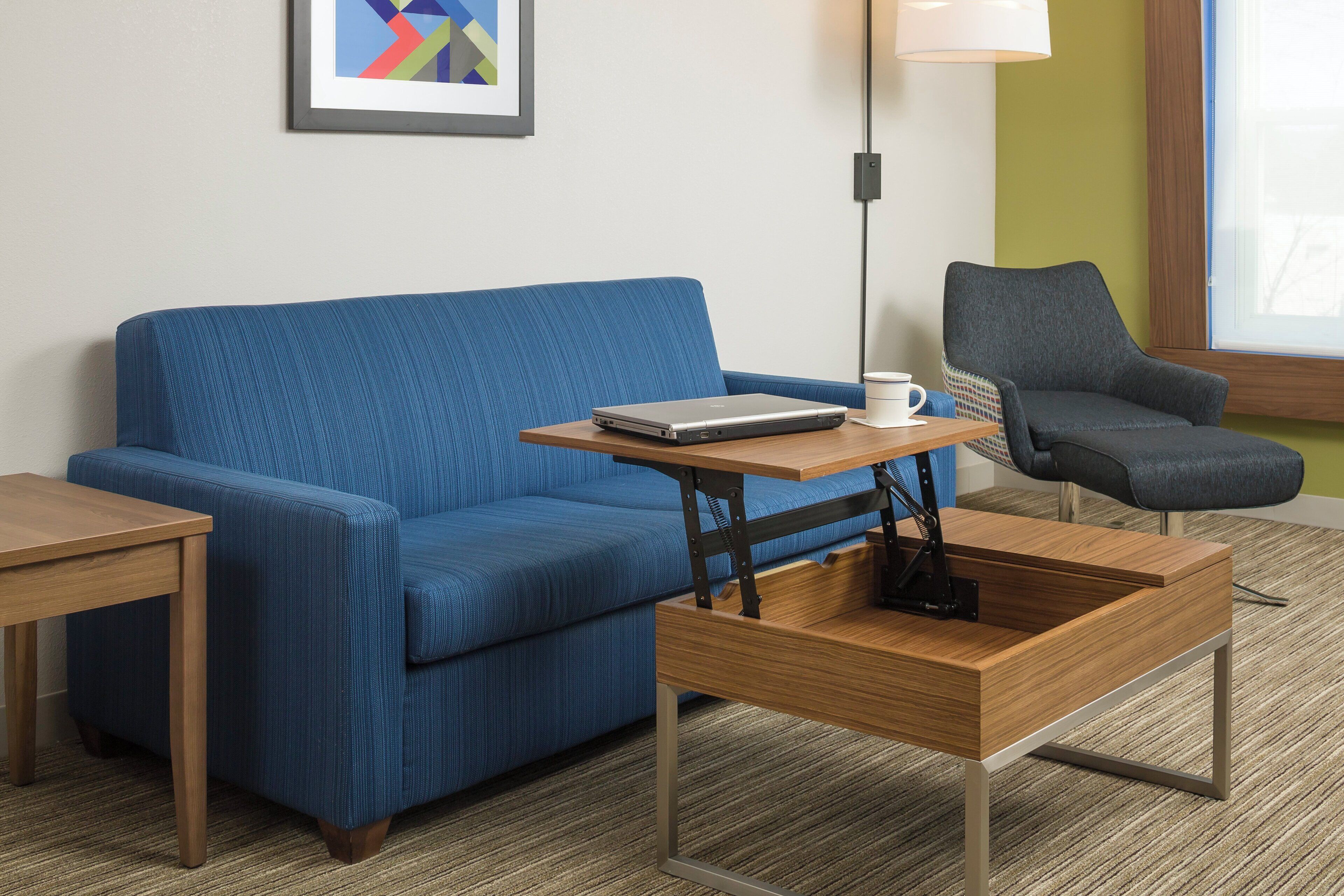 Una habitación moderna con un sofá azul, una mesa de centro de madera, una computadora portátil, una taza blanca y un sillón gris.