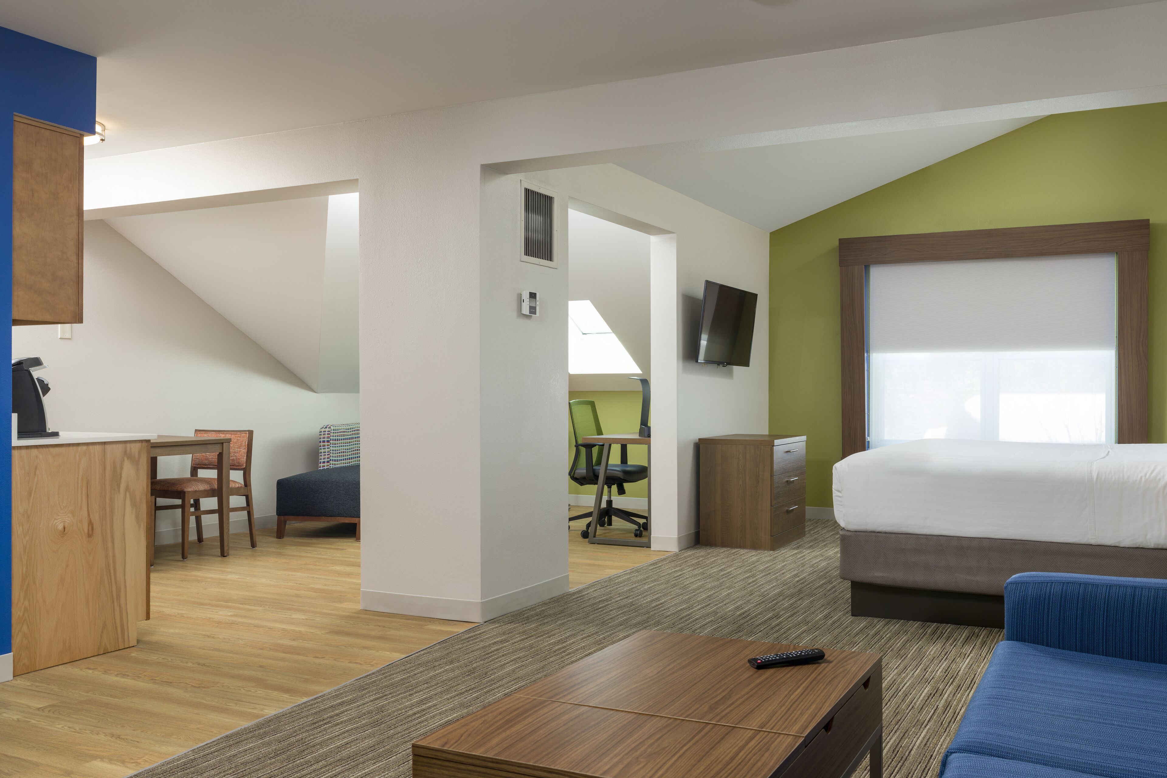 A modern hotel room with a large bed, a desk, and a chair. There is a TV mounted on the wall and a window providing natural light.