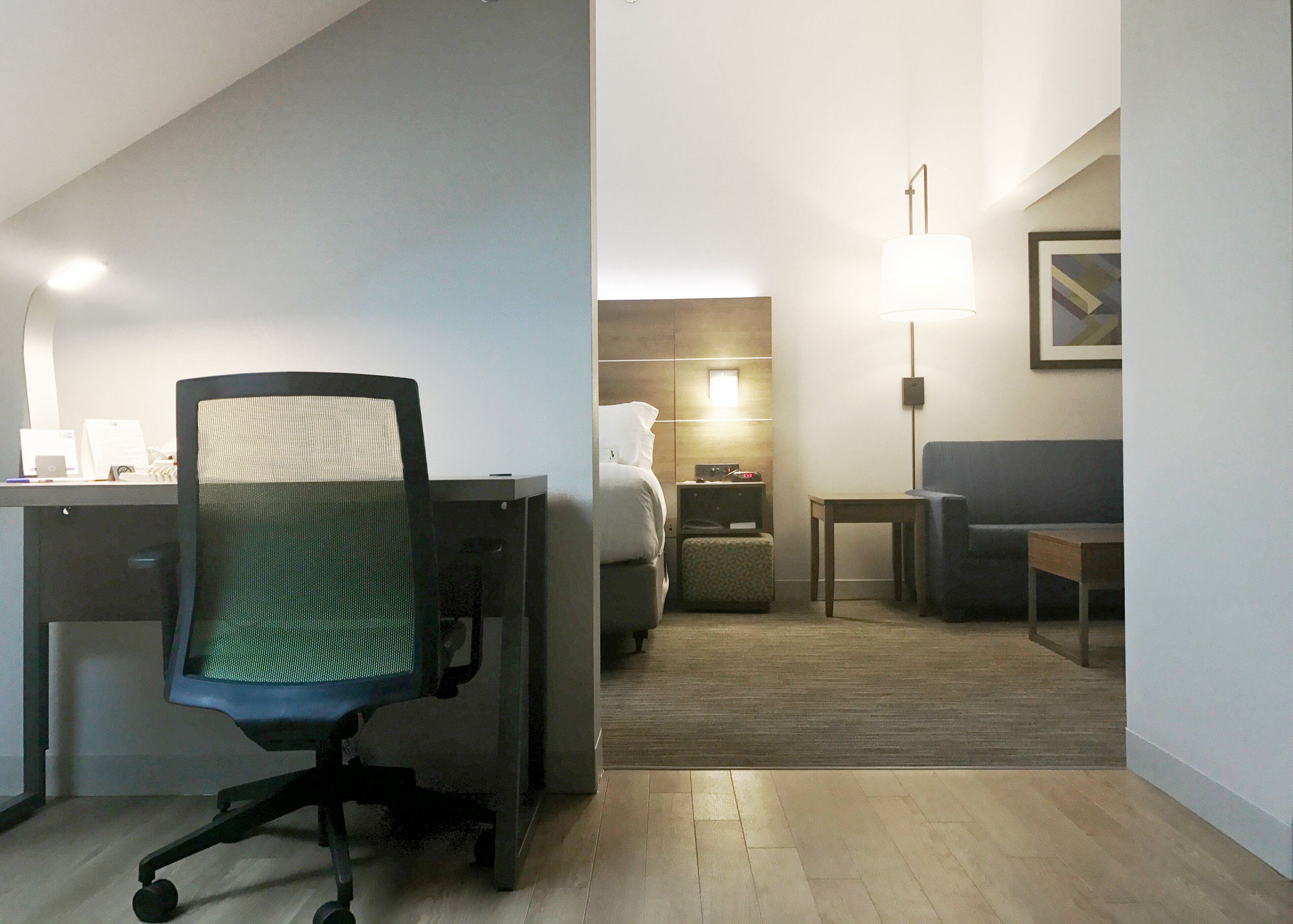 Una habitación de hotel con escritorio, silla de oficina, cama, mesita de noche, lámpara y un cuadro en la pared.