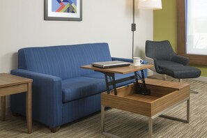 Ein modernes Hotelzimmer mit einem blauen Sofa, einem Holztisch, einem Laptop, einer weißen Tasse und einem grauen Sessel.