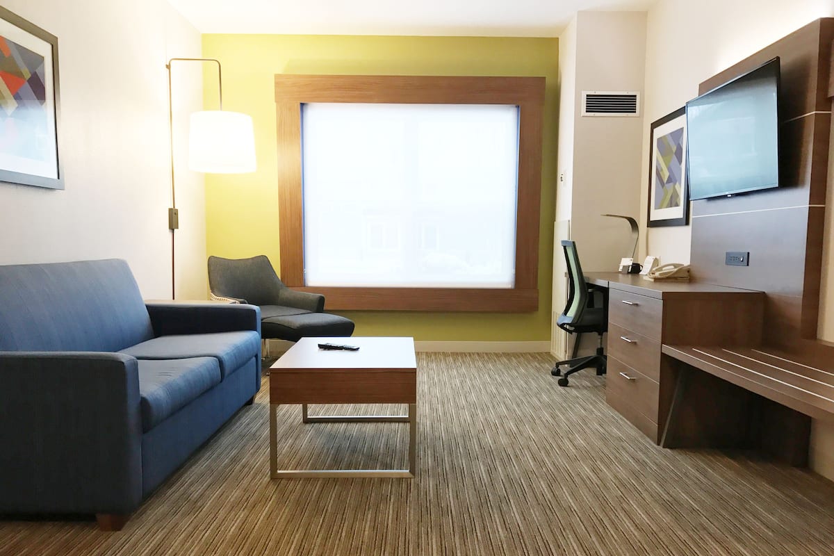 Une chambre d’hôtel avec un canapé bleu, un bureau en bois, une télévision et une fenêtre avec des stores.