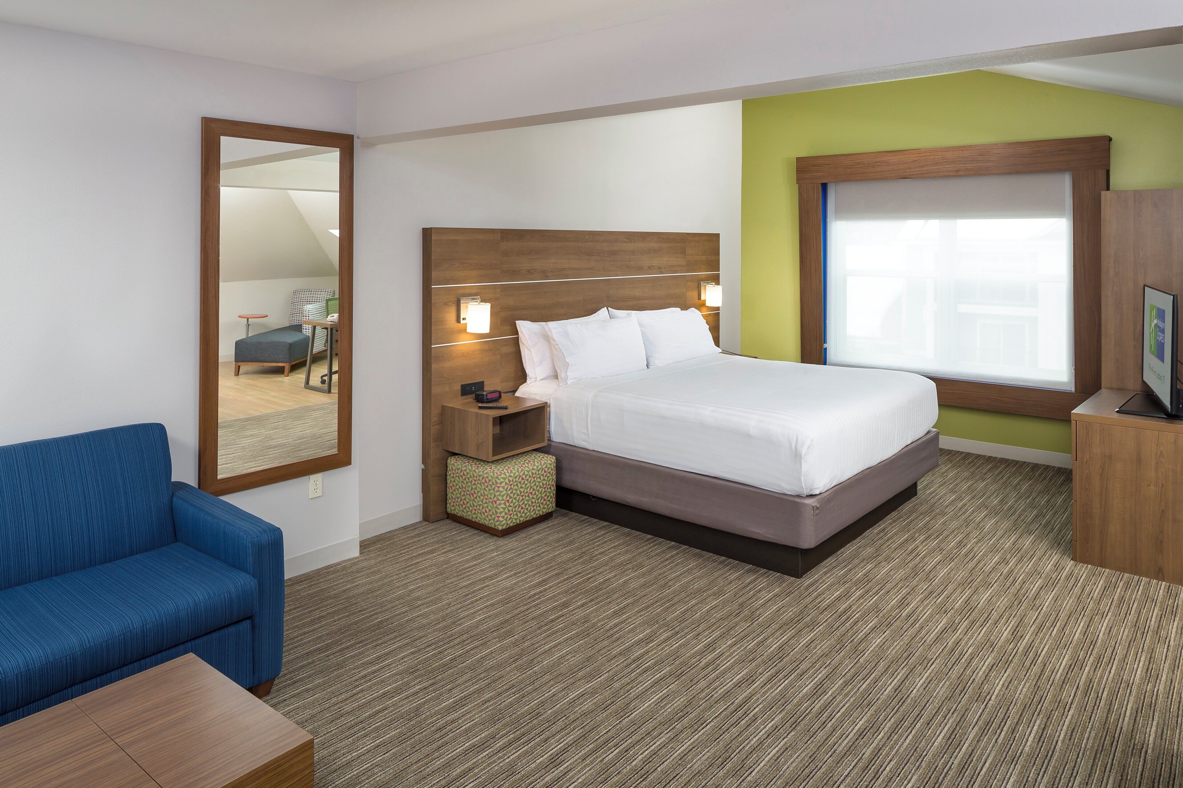 A hotel room with a bed, a desk, a chair, a nightstand, a mirror, and a window.