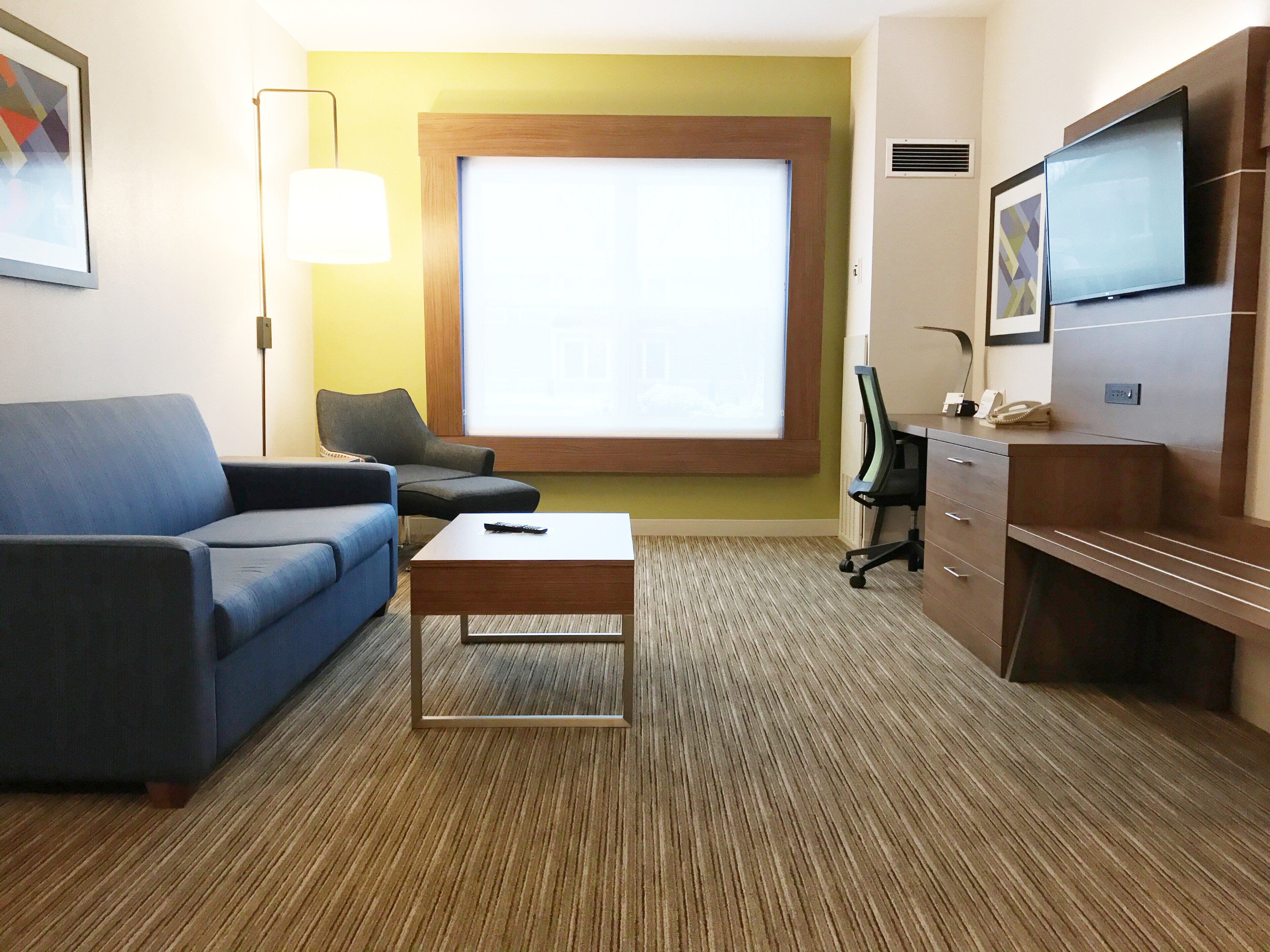 Habitación de hotel con un sofá azul, un escritorio de madera, un televisor y un ventanal grande.