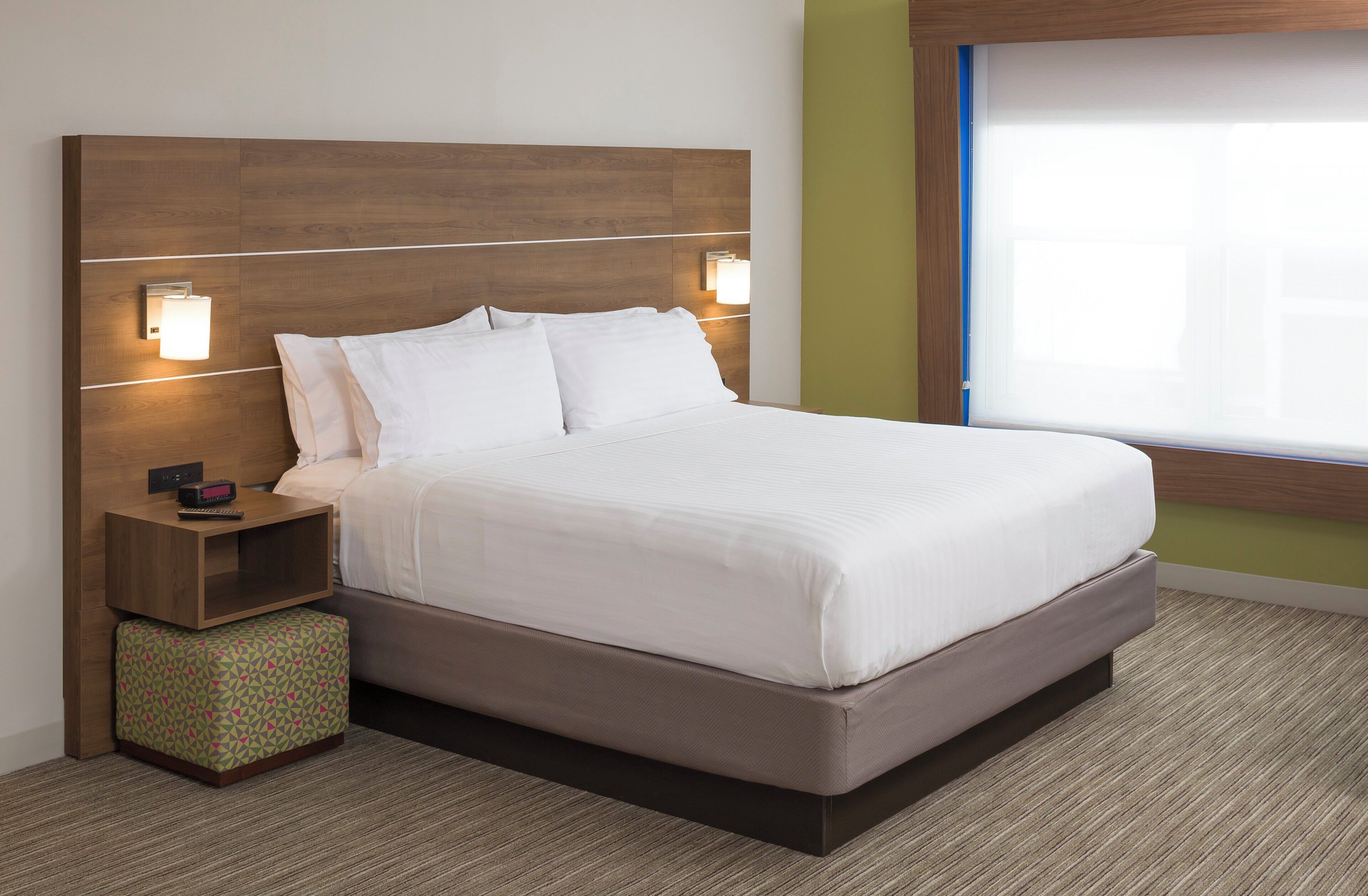 Quarto de hotel com uma cama grande, duas luminárias de cabeceira, uma mesa de cabeceira com um rádio e uma janela com persianas.