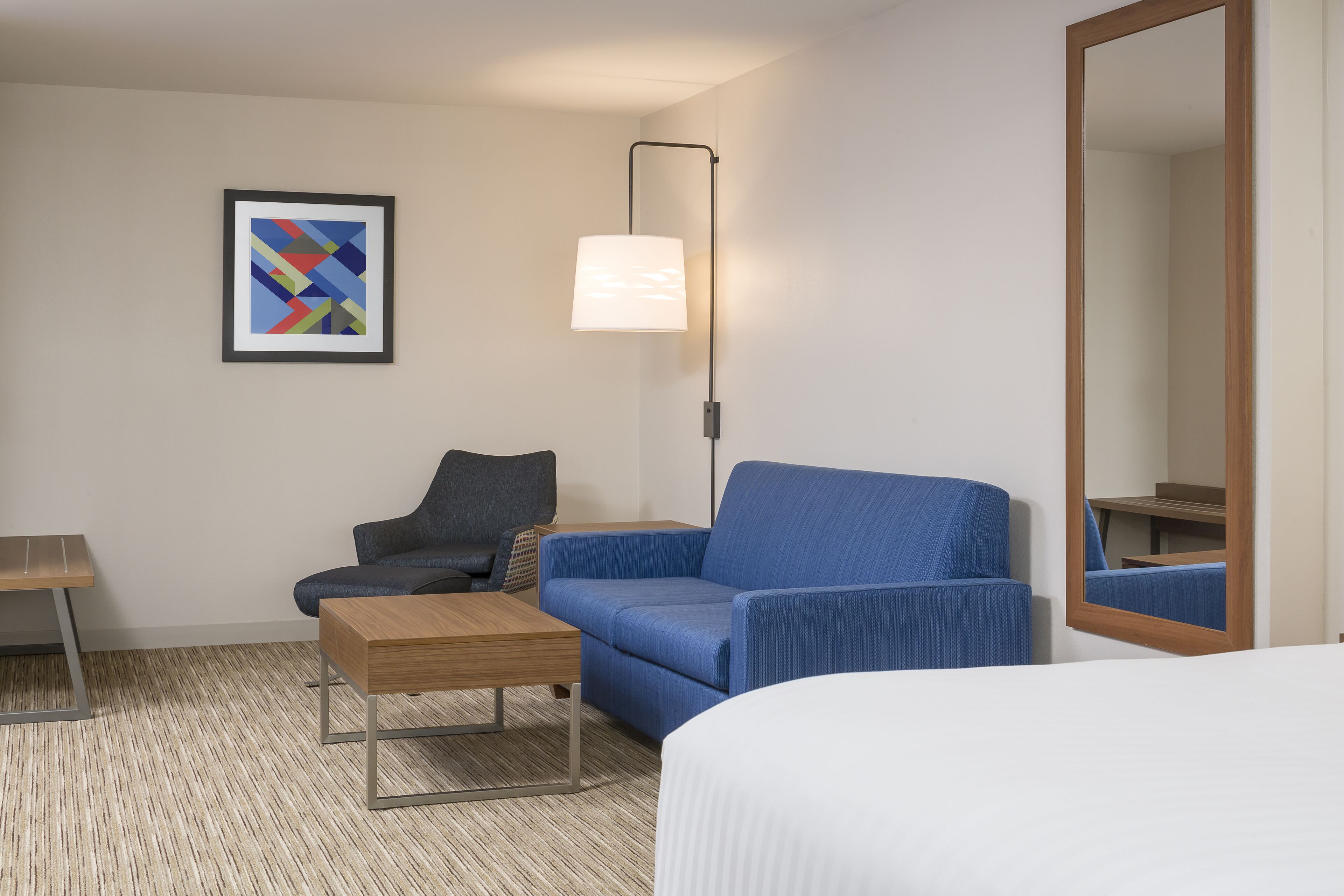 Ein Hotelzimmer mit einem Bett, einem blauen Sofa, einem Holznachttisch, einem kleinen Schreibtisch, einer Lampe, einem gerahmten abstrakten Gemälde und einem Spiegel.