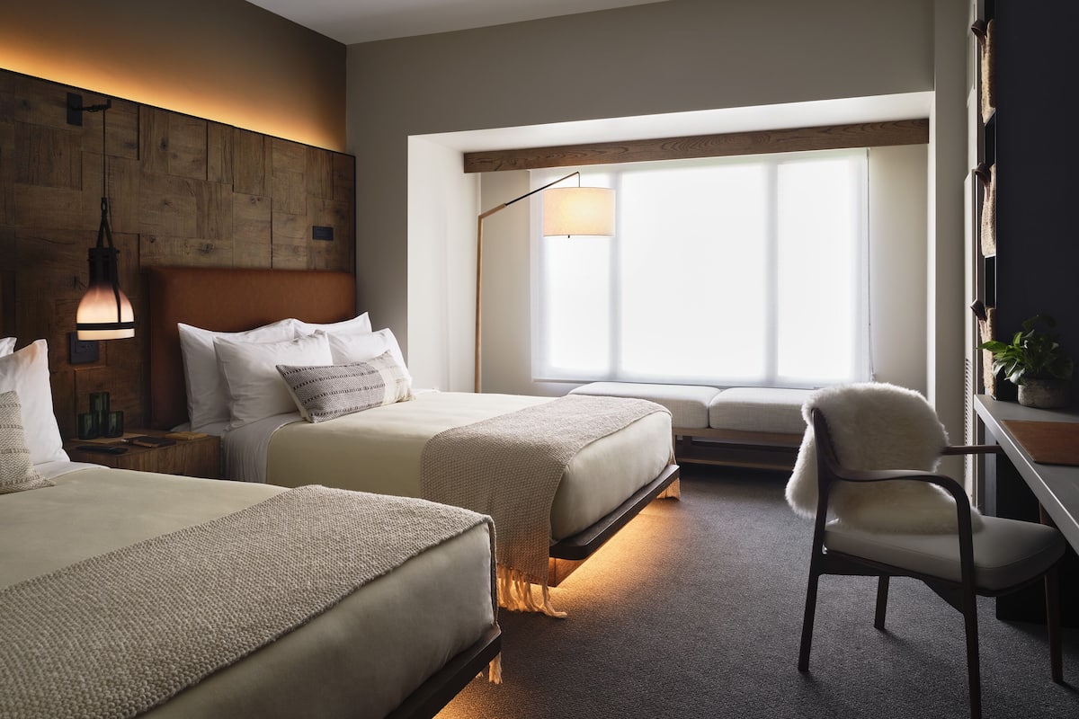 Ein Hotelzimmer mit zwei Betten, einem Schreibtisch, einem Stuhl, einem großen Fenster und einer Wandlampe.