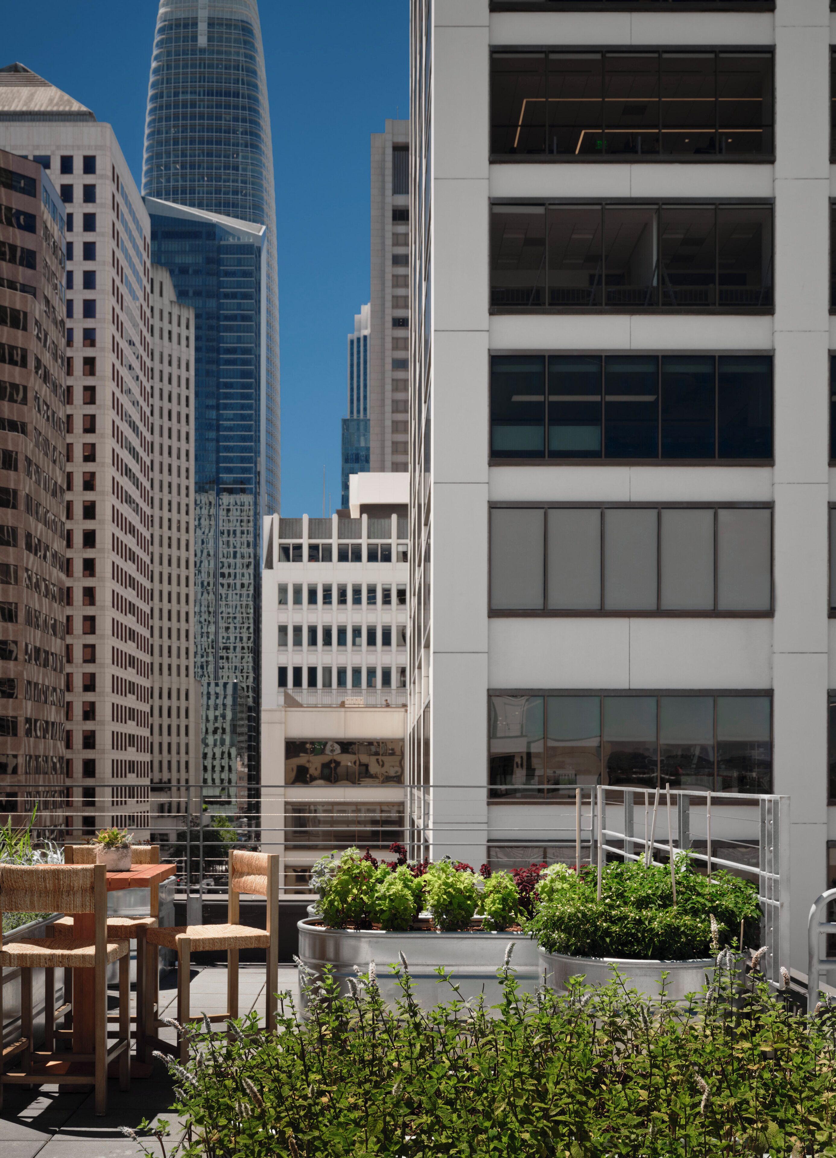 A rooftop terrace with outdoor seating and planters, surrounded by modern buildings.