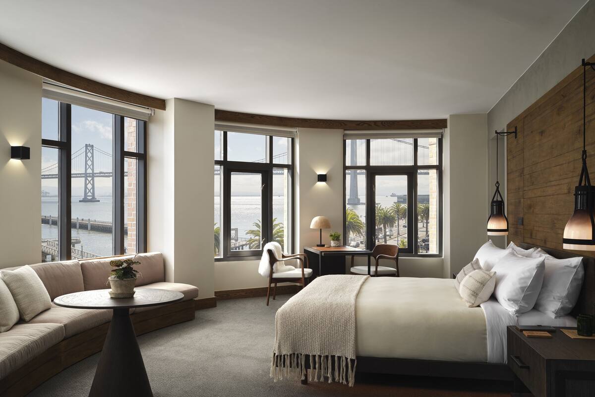 Ein modernes Hotelzimmer mit einer geschwungenen Gestaltung, großen Fenstern mit Blick auf eine Brücke und den Wasserblick, einer gemütlichen Sitzecke und einem ordentlich bezogenen Bett.