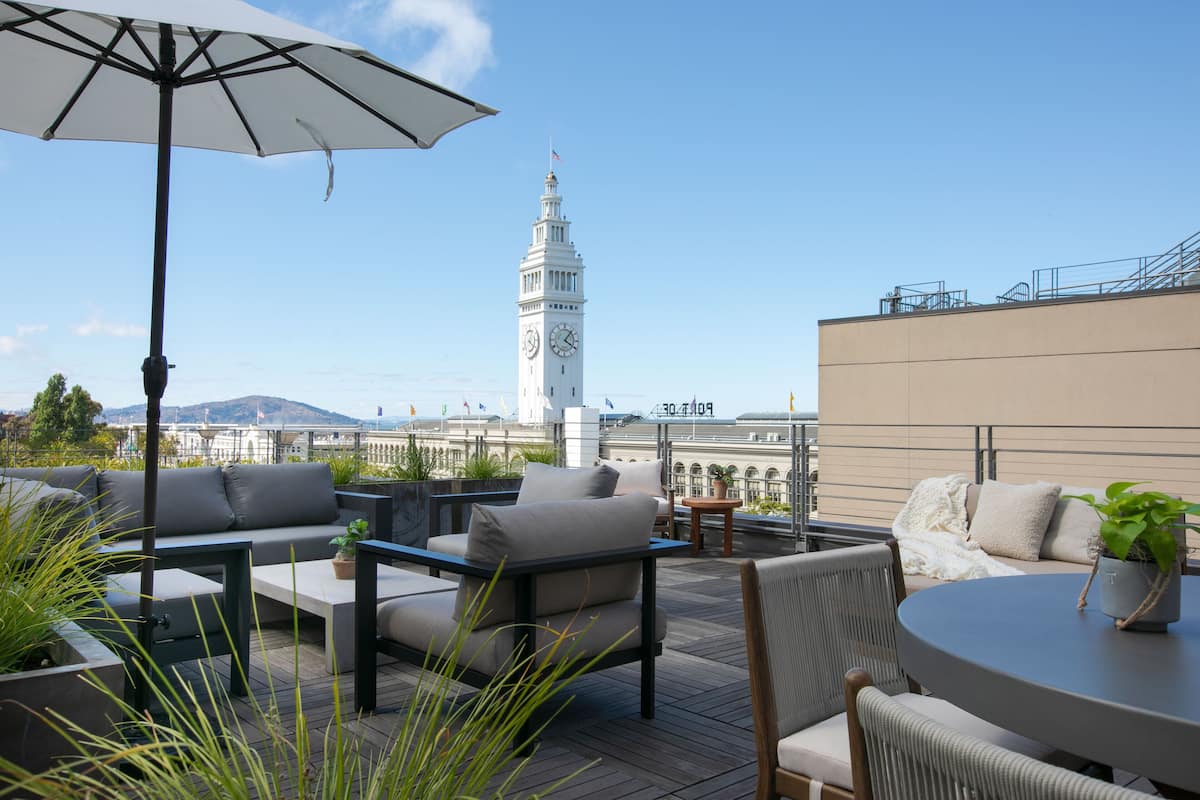 Eine Dachterrasse mit Außenmöbeln, im Hintergrund ein Glockenturm und ein klarer Himmel.