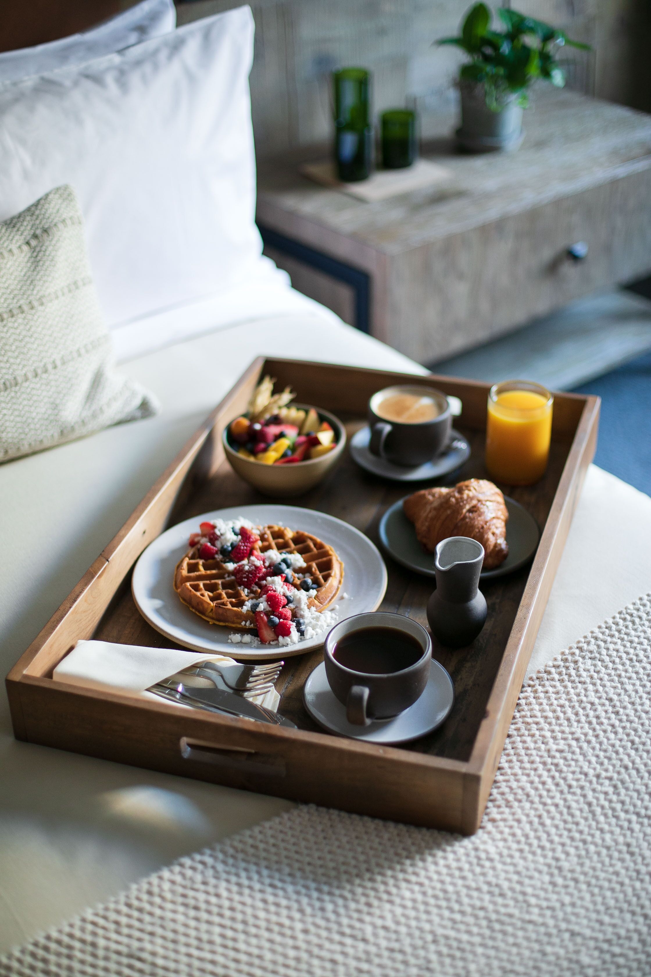 A breakfast tray on a bed with waffles, croissants, fruit, coffee, and juice.