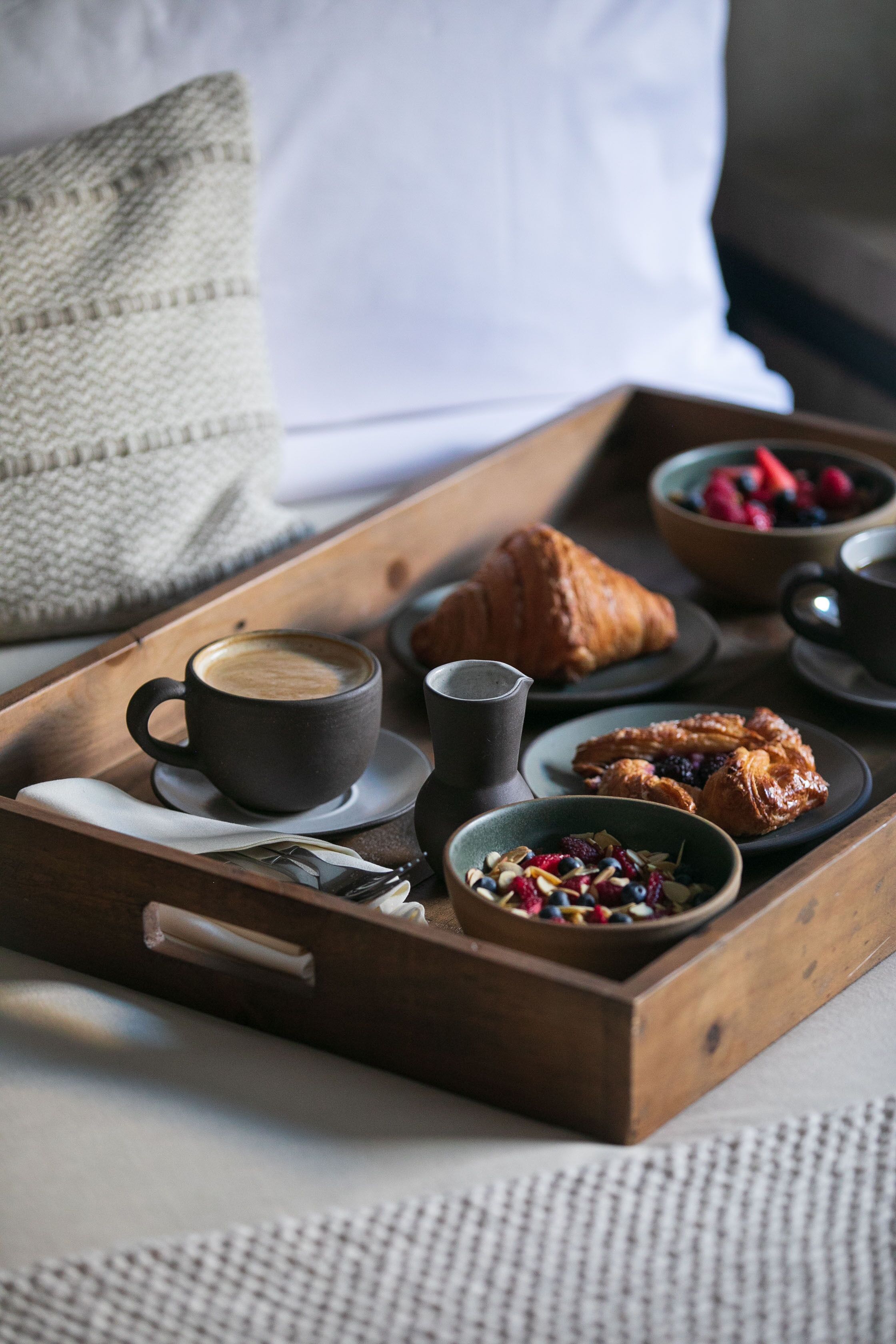 A wooden tray with a cup of coffee, a croissant, and bowls of berries and nuts.