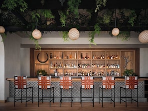 Un espace bar avec des étagères en bois remplies de bouteilles, une décoration murale incurvée, des plantes suspendues et des tabourets de bar avec des dossiers en osier tressé.