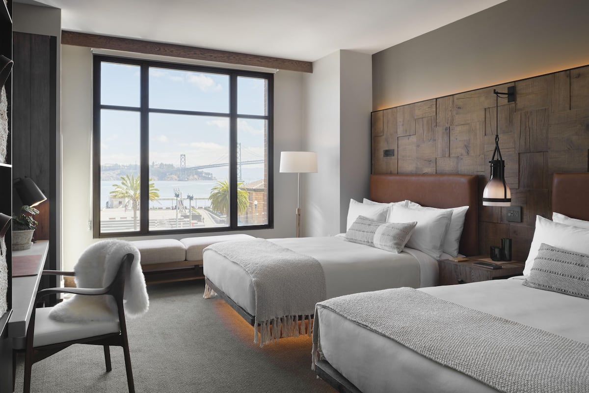 Ein Hotelzimmer mit zwei Betten, einem großen Fenster mit Blick auf eine Brücke, einem Schreibtisch mit Stuhl und einem Nachttisch mit Lampe.