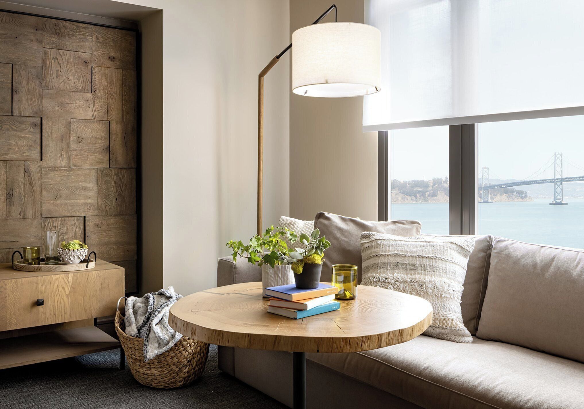 A modern living room with a round wooden table, a sofa, a floor lamp, and a view of a bridge.
