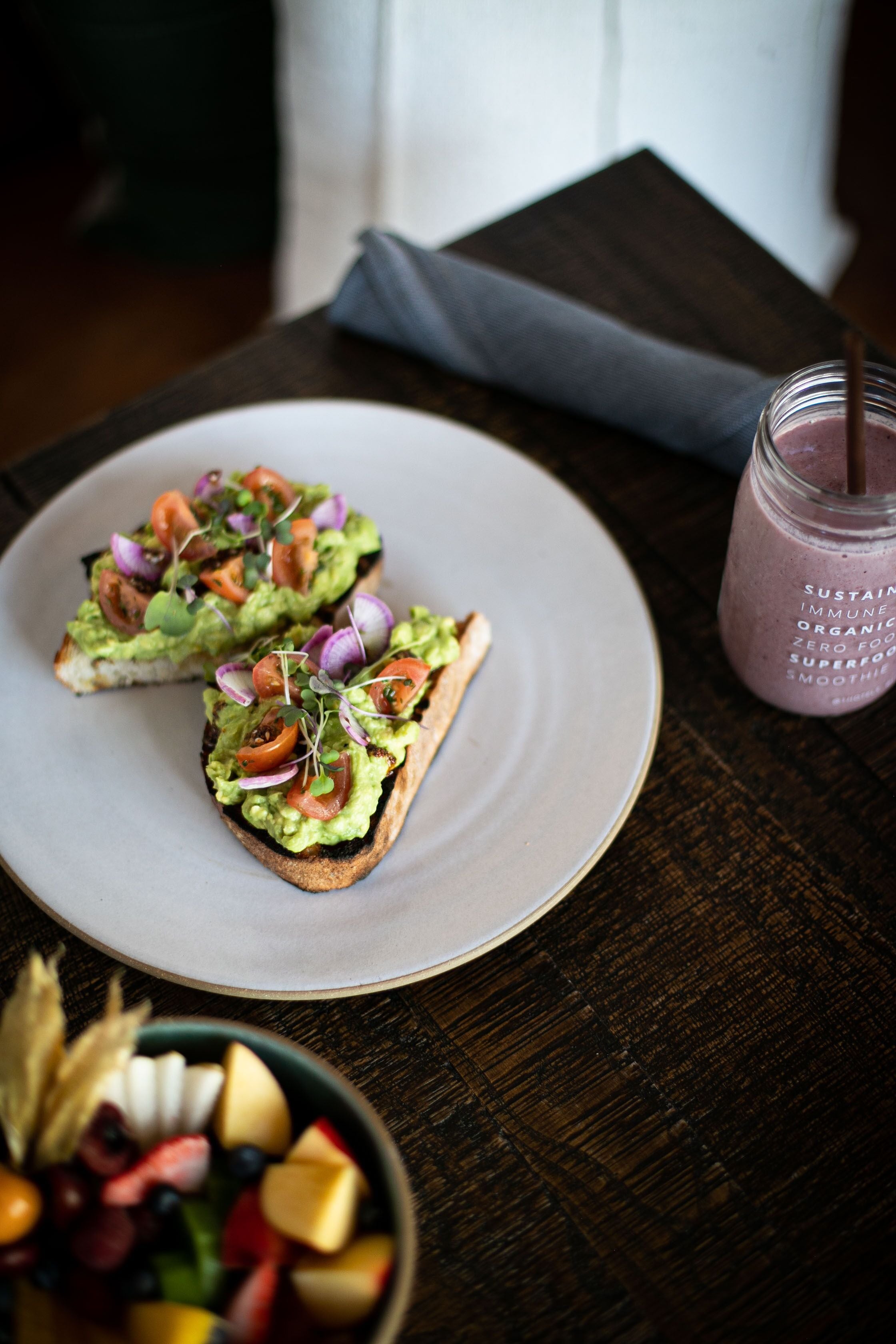 Two avocado toast sandwiches on a white plate, a glass of smoothie, and a bowl of fruit on a wooden table.