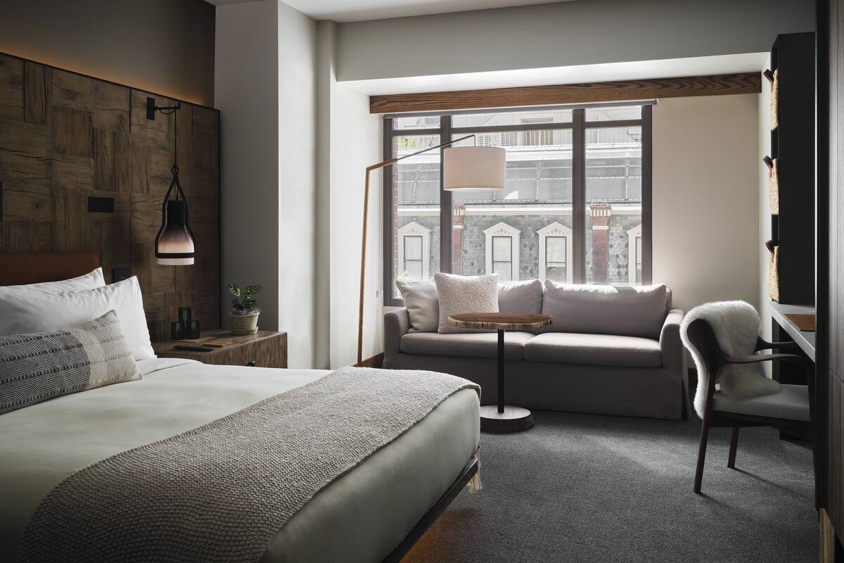 Ein modernes Hotelzimmer mit einem großen Bett, einem Sofa, einem Sessel und einem Fenster mit Blick auf Gebäude.