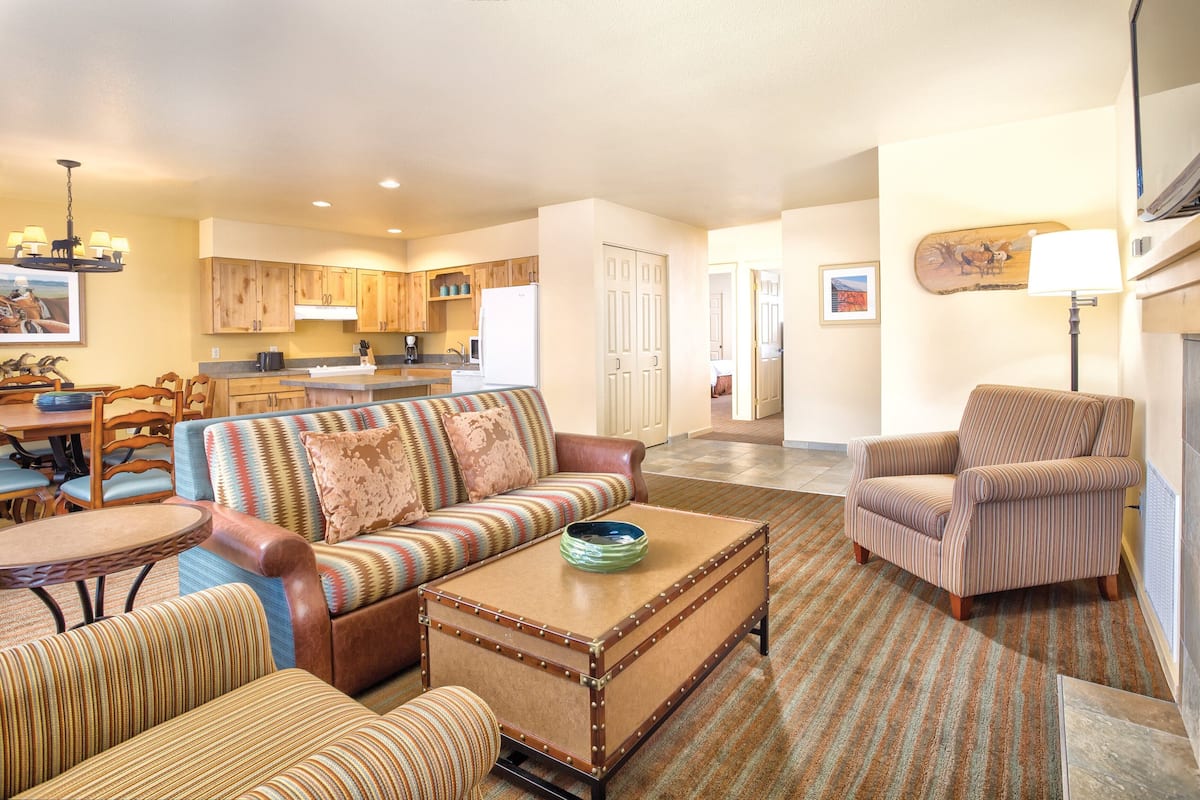 A spacious living room with a sofa, armchairs, and a coffee table. There is a kitchen in the background with wooden cabinets and a refrigerator.