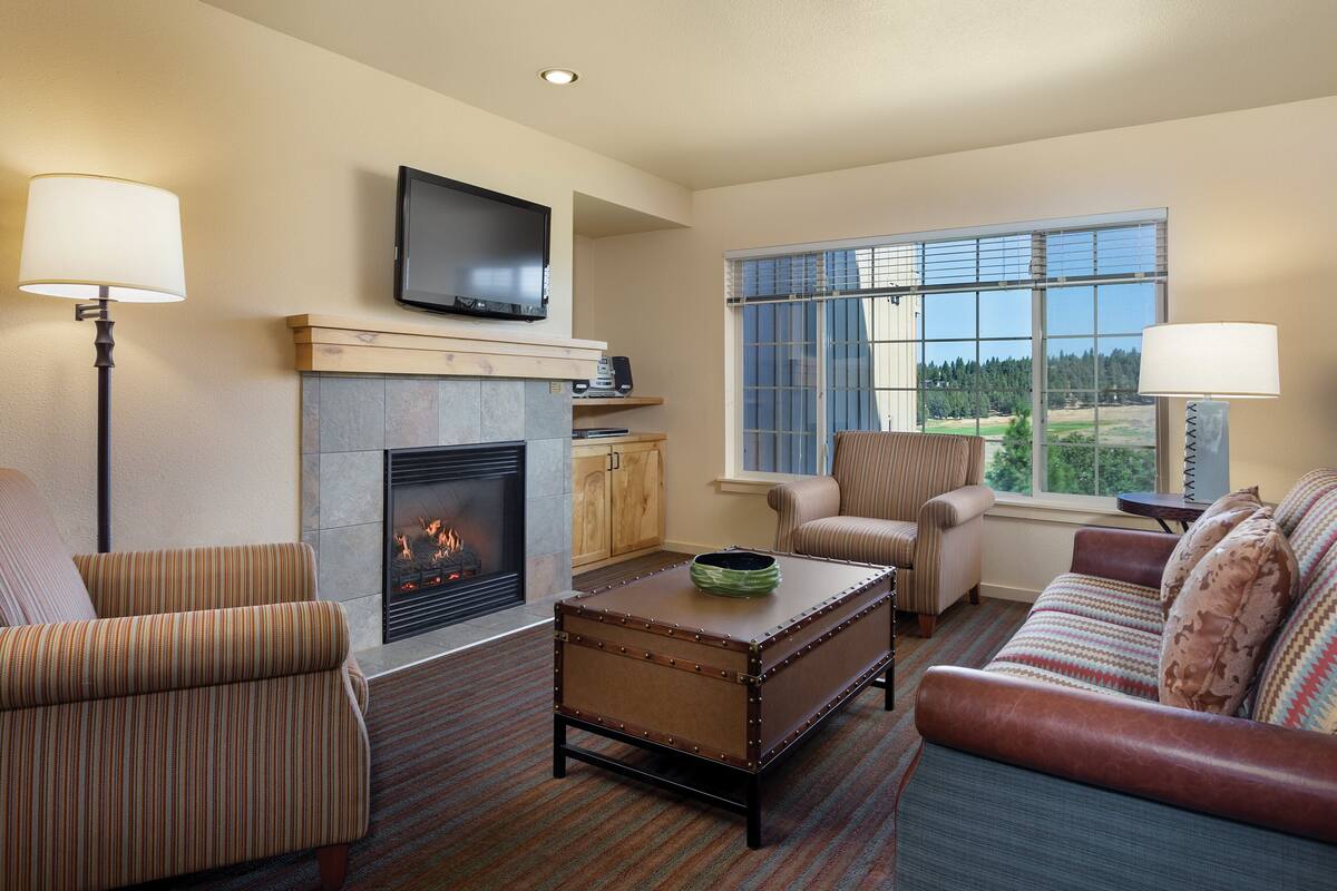 A living room with a fireplace, a television mounted above it, and a large window offering a view of the outdoors.